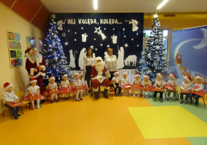 Pani dyrektor Maria Królikowska, pani Ewelina Cicha i dzieci zebrane wokół Mikołaja pozują na tle dekoracji świątecznej.